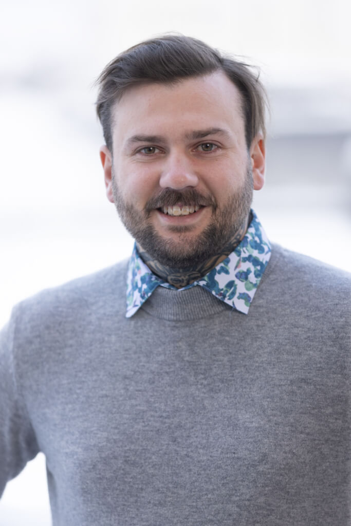 Mann mit grauem Pullover und bunter Bluse, lächelt in die Kamera.