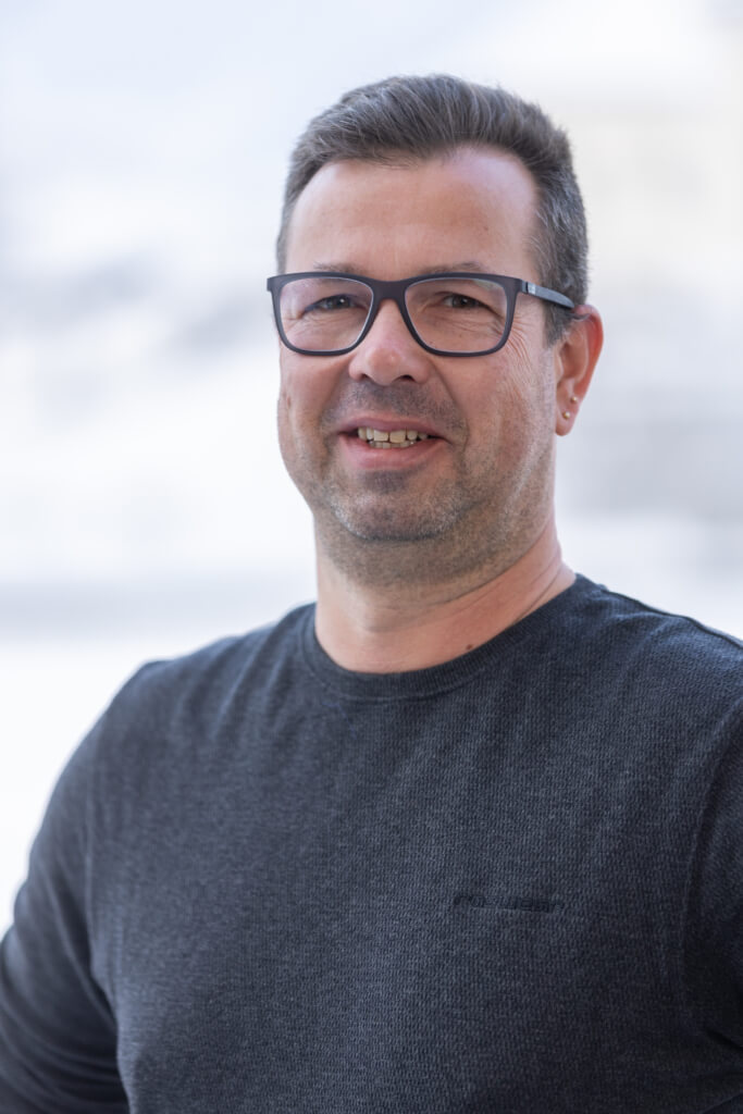 Mann in dunklem Shirt mit Brille, freundlich lächelnd, vor unscharfem Hintergrund.