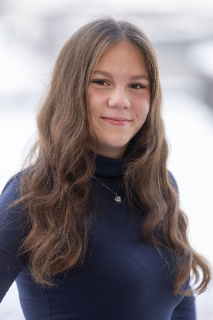 Junge Frau mit langen, lockigen Haaren in einem blauen Rollkragenpullover, lächelt freundlich.