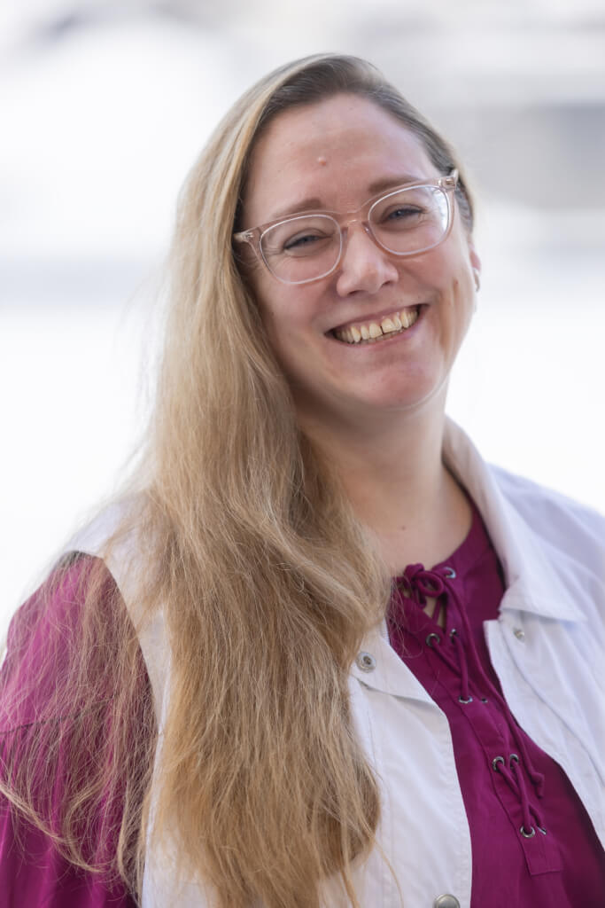 Lächelnde Frau mit langen Haaren, im lila Oberteil und weißer Jacke, vor hellem Hintergrund.