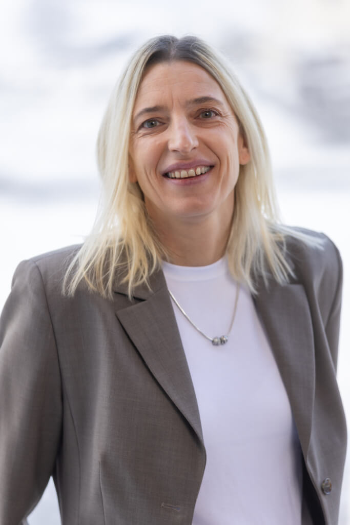 Lächelnde Person mit blonder Haarfarbe in einem grauen Blazer, Hintergrund unscharf.