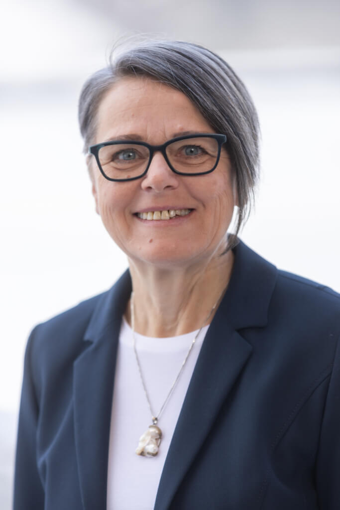 Portrait einer engagierten Frau in dunklem Blazer, lächelnd, vor hellem Hintergrund.