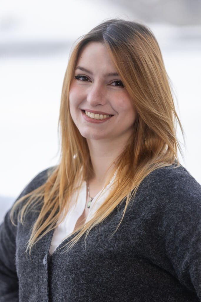 Lächelnde Frau mit langen, blonden Haaren in grauem Pullover, symbolisiert Gemeinschaft und Engagement.