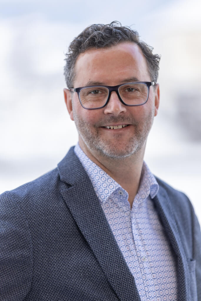 Porträt eines Mannes in Anzug mit Brille, lächelnd, Hintergrund unscharf.